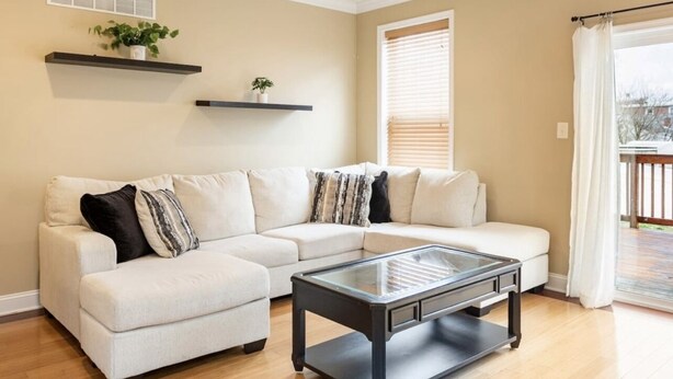 A living room with a sectional sofa, a glass coffee table, and a window with blinds. - Charming 3BR & 2.5Ba nested in KOP w free parking, close to Conshy & Philly. (Bridgeport)