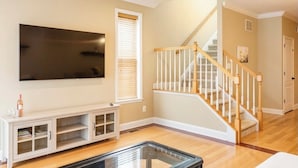 A living room with a flat-screen TV mounted on the wall, a wooden staircase, and a white entertainment unit. - Charming 3BR & 2.5Ba nested in KOP w free parking, close to Conshy & Philly. (Bridgeport)