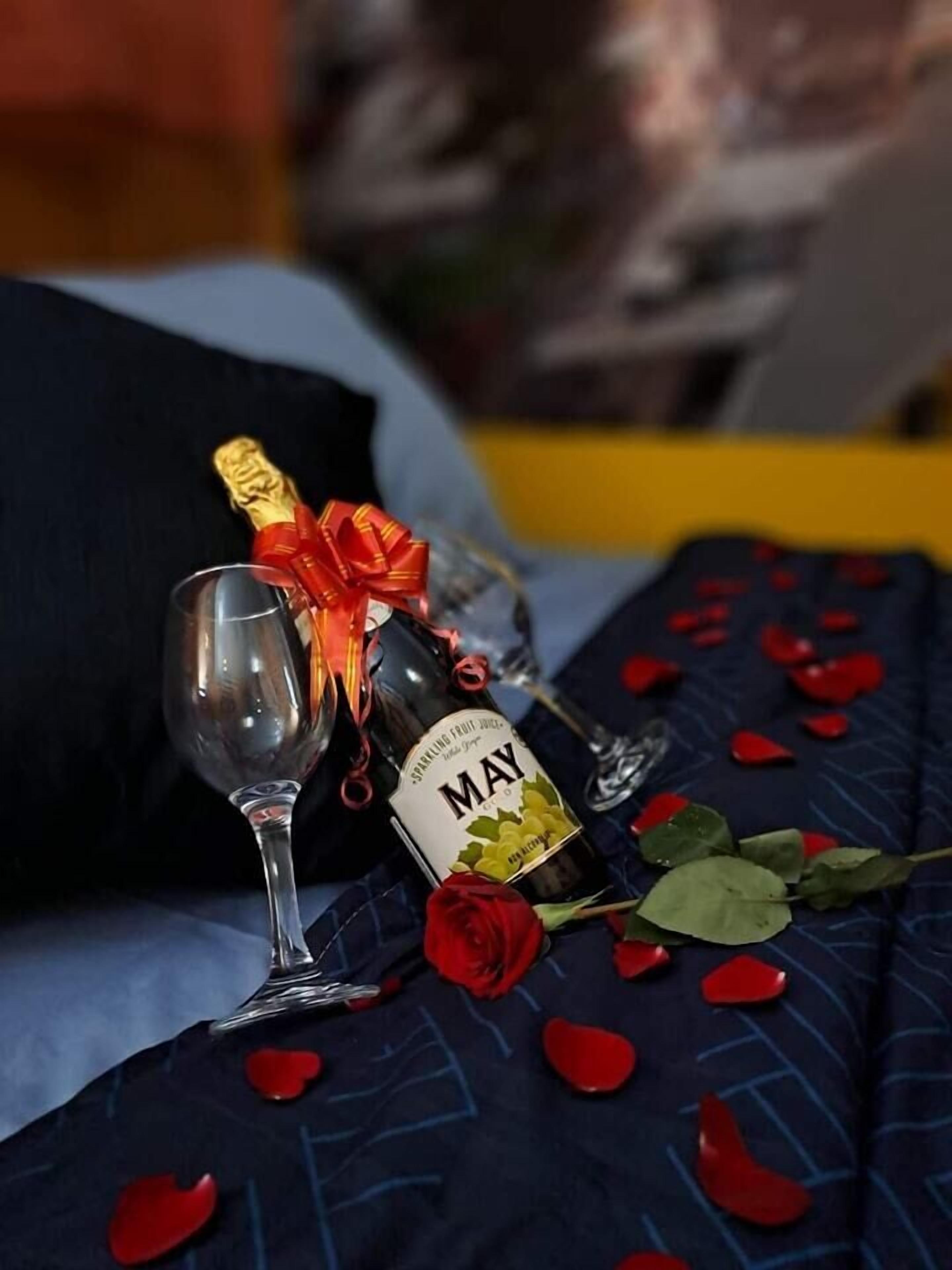 Un lit recouvert d’une couverture bleu foncé, ornée de pétales de roses rouges et surmonté d’une seule rose rouge. Une bouteille de jus de fruits pétillant de la marque May et un verre à vin sont posés sur le lit.