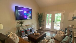 A living room with a brown leather couch, a flat-screen TV mounted on the wall, and a sliding glass door leading to an outdoor area. - Cozy and Relaxing Lake House close to Lake of the Arbuckles (Sulphur)