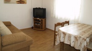 A living room with a sofa, a TV on a wooden stand, a dining table with chairs, and a window with curtains. - Stunning sea View From one Bedroom Apartment Reno (Preko)