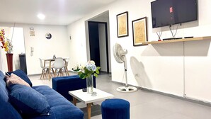 A modern living room with a sofa, a small table with a vase, a standing fan, and a TV mounted on the wall. - Apto Super Cómodo al Norte de la Ciudad (Valledupar)