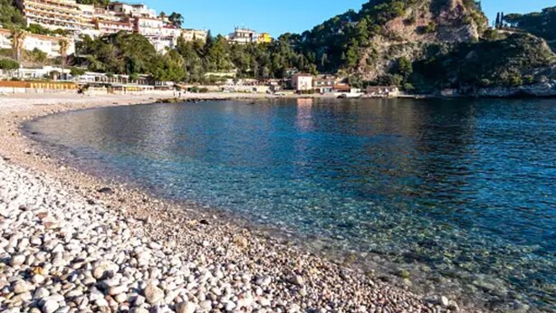 A pebble beach with clear water, buildings on a hillside, and a rocky cliff. - Fabulous Villa in Albarella Private Island by Beahost Rentals (Rosolina)
