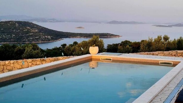 Una piscina rectangular con borde de piedra, rodeada de vegetación y con vistas a una masa de agua y tierras lejanas.