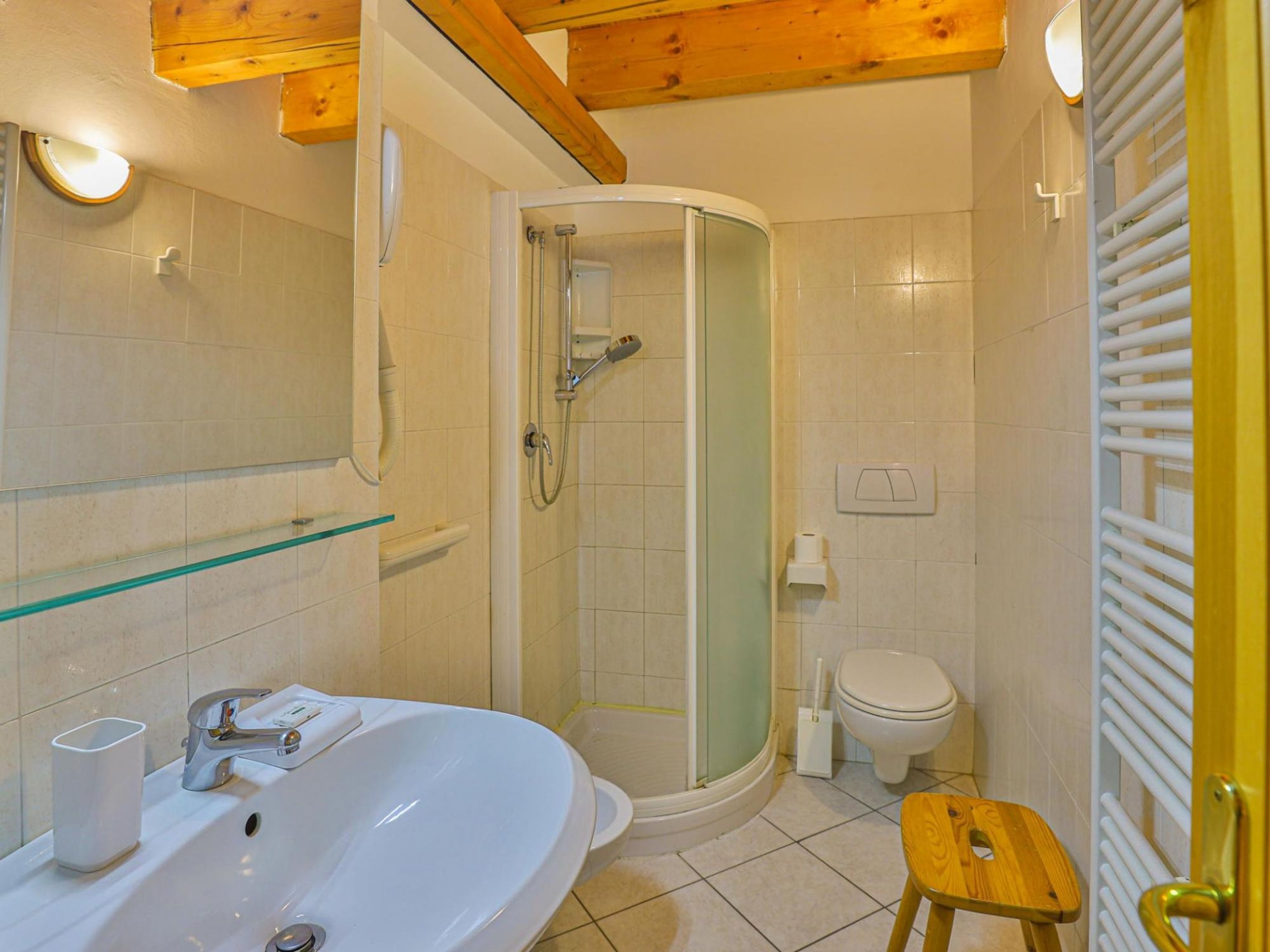 A modern bathroom with a glass-enclosed shower, a white sink with a mirror, and a wooden stool.