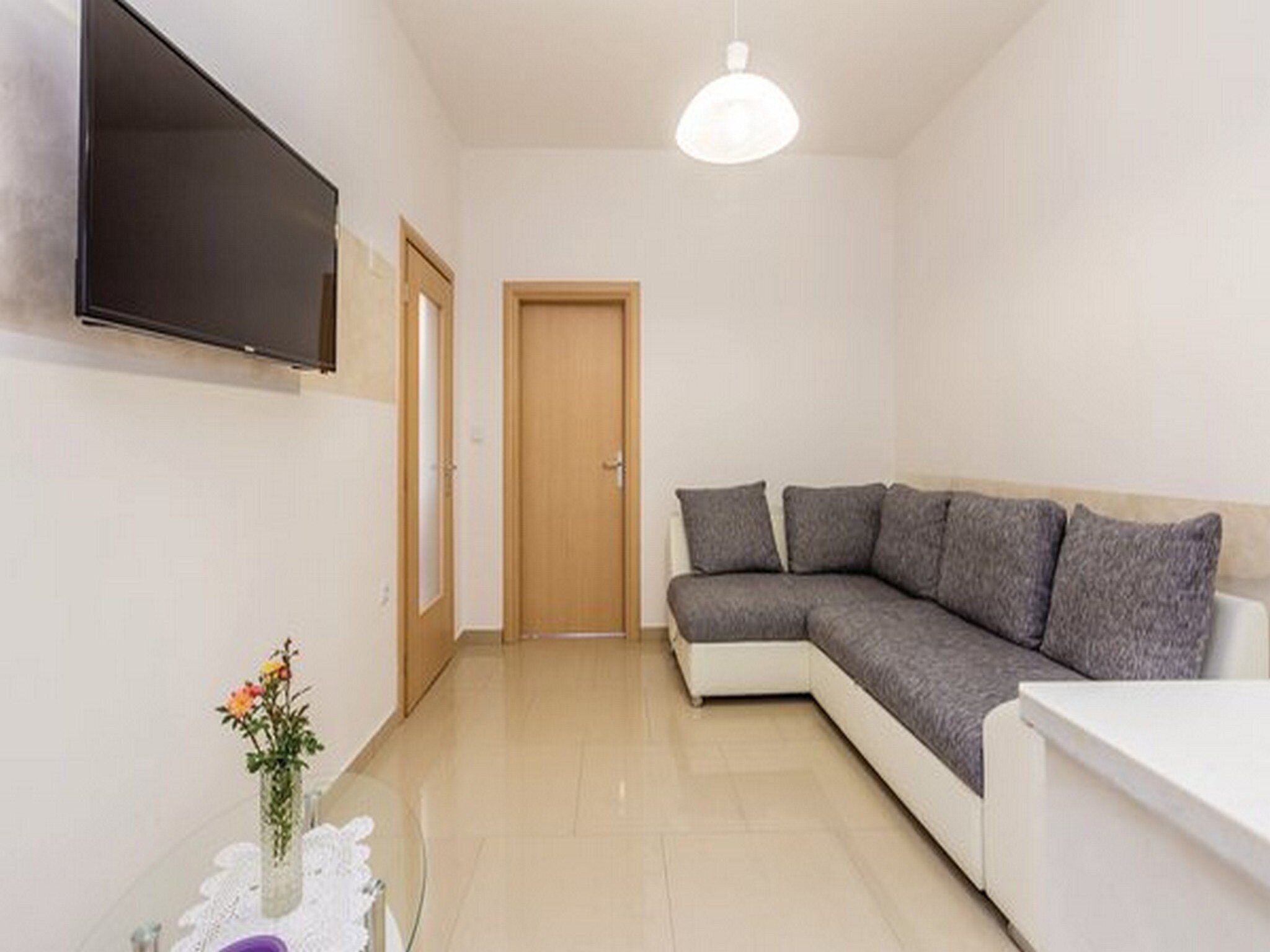 A modern living room with a flat-screen TV mounted on the wall, a wooden door, a grey sectional sofa, and a small table with a vase of flowers.