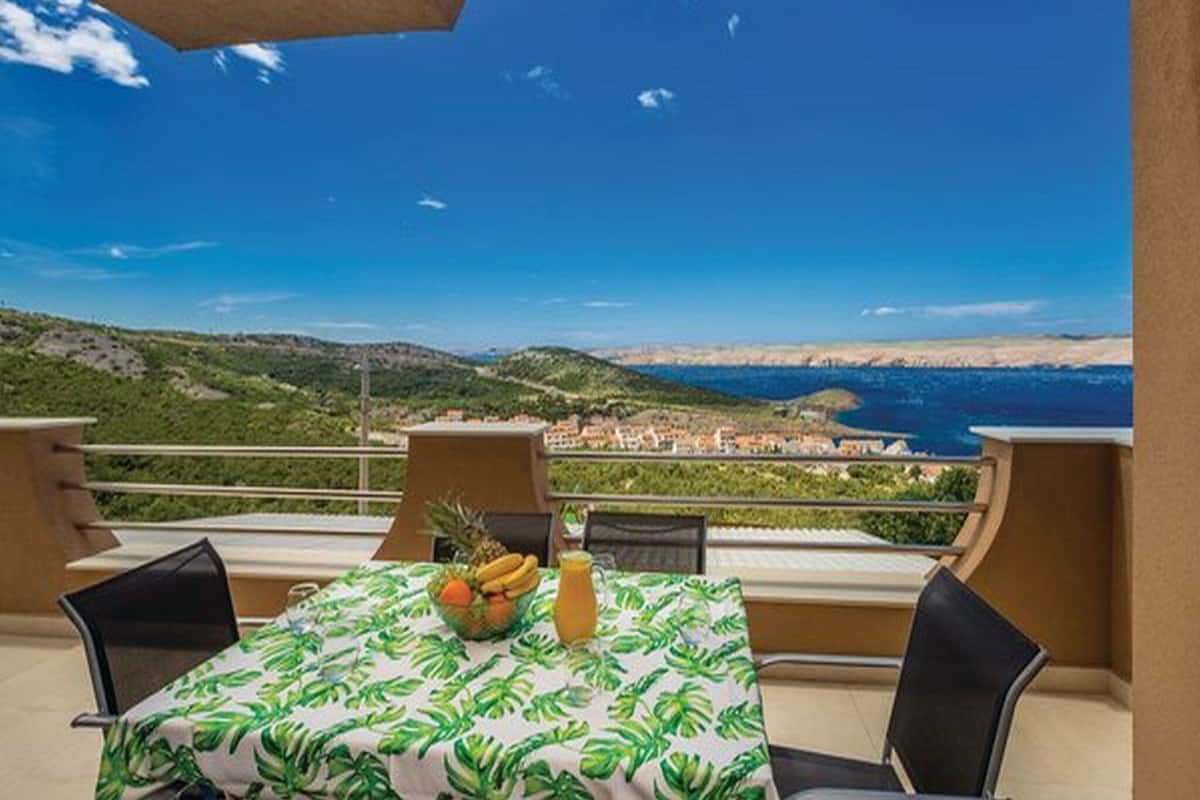 A table set outdoors with a tropical tablecloth, a fruit bowl, and a drink, overlooking a coastal landscape with hills and buildings.