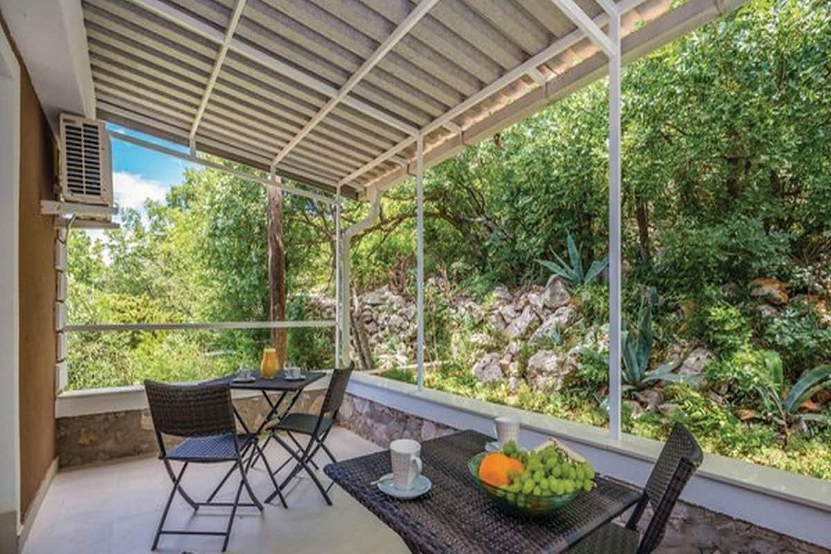 A covered outdoor seating area with wicker furniture, a table with a fruit bowl, and a view of lush greenery and rocks.