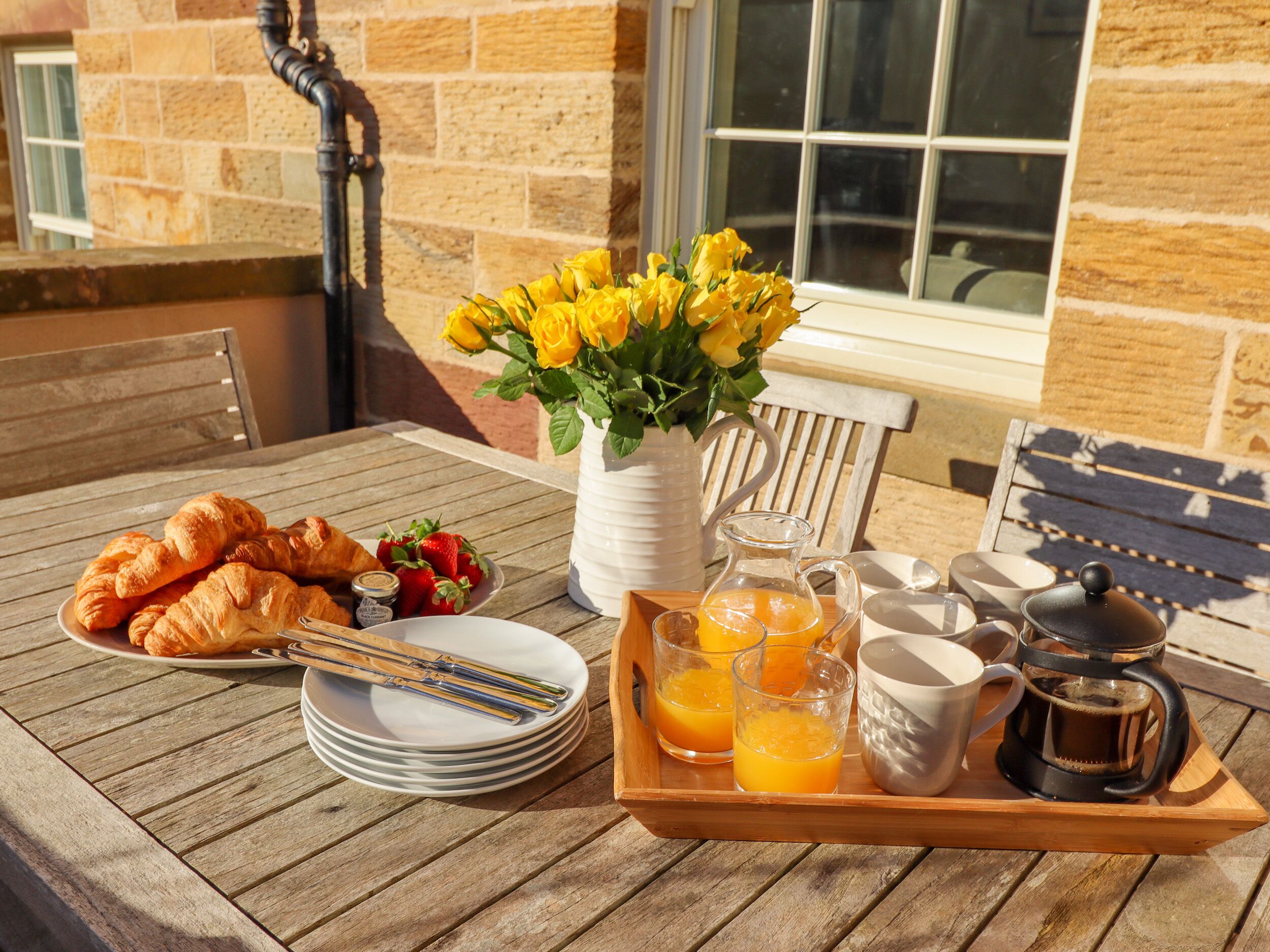 A breakfast setup on an outdoor table with croissants, strawberries, a pitcher of orange juice, and a coffee pot.