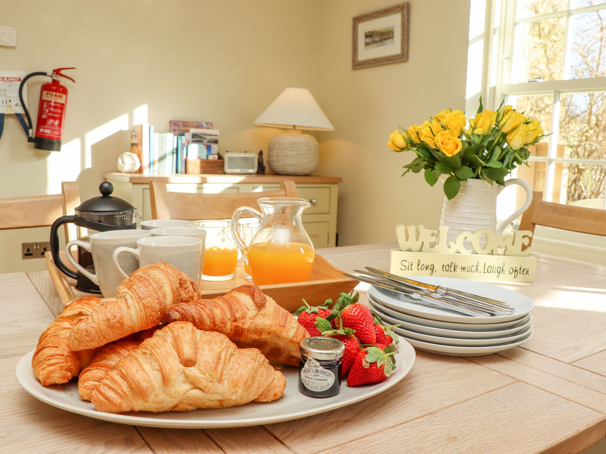 Een ontbijttafel met croissants, aardbeien en kruisbessenjam op een bord, een karaf sinaasappelsap en een koffiepot op een houten tafel.