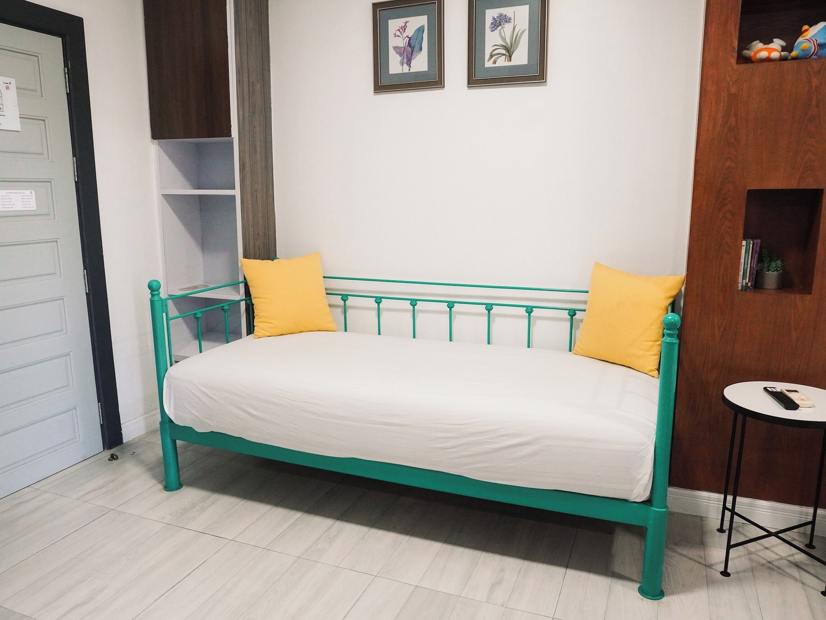 A teal metal bed with white bedding, two yellow pillows, a small round table with a remote, and a shelf with books and a plant.