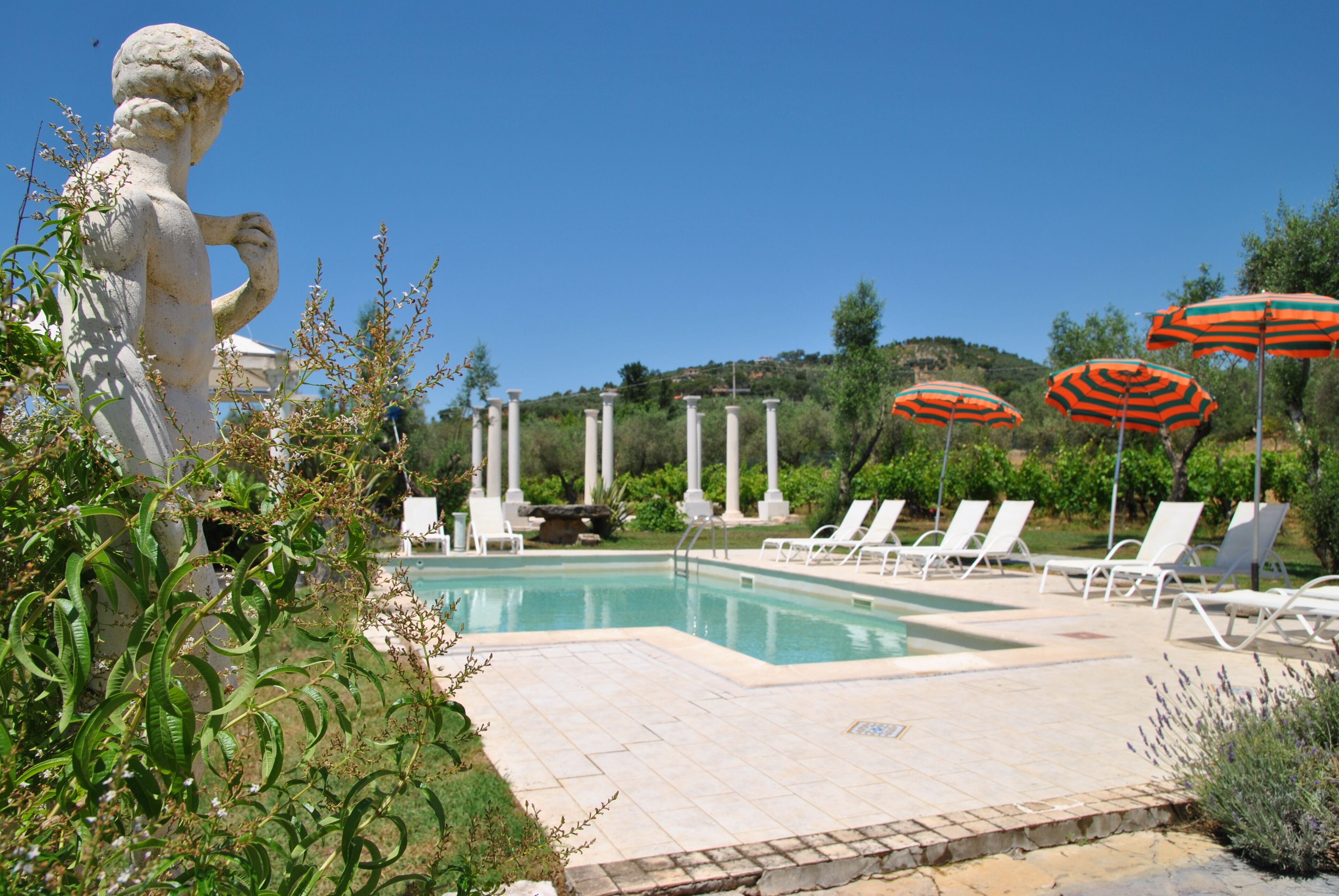 A pool area with white lounge chairs and orange striped umbrellas, surrounded by greenery and a statue.
