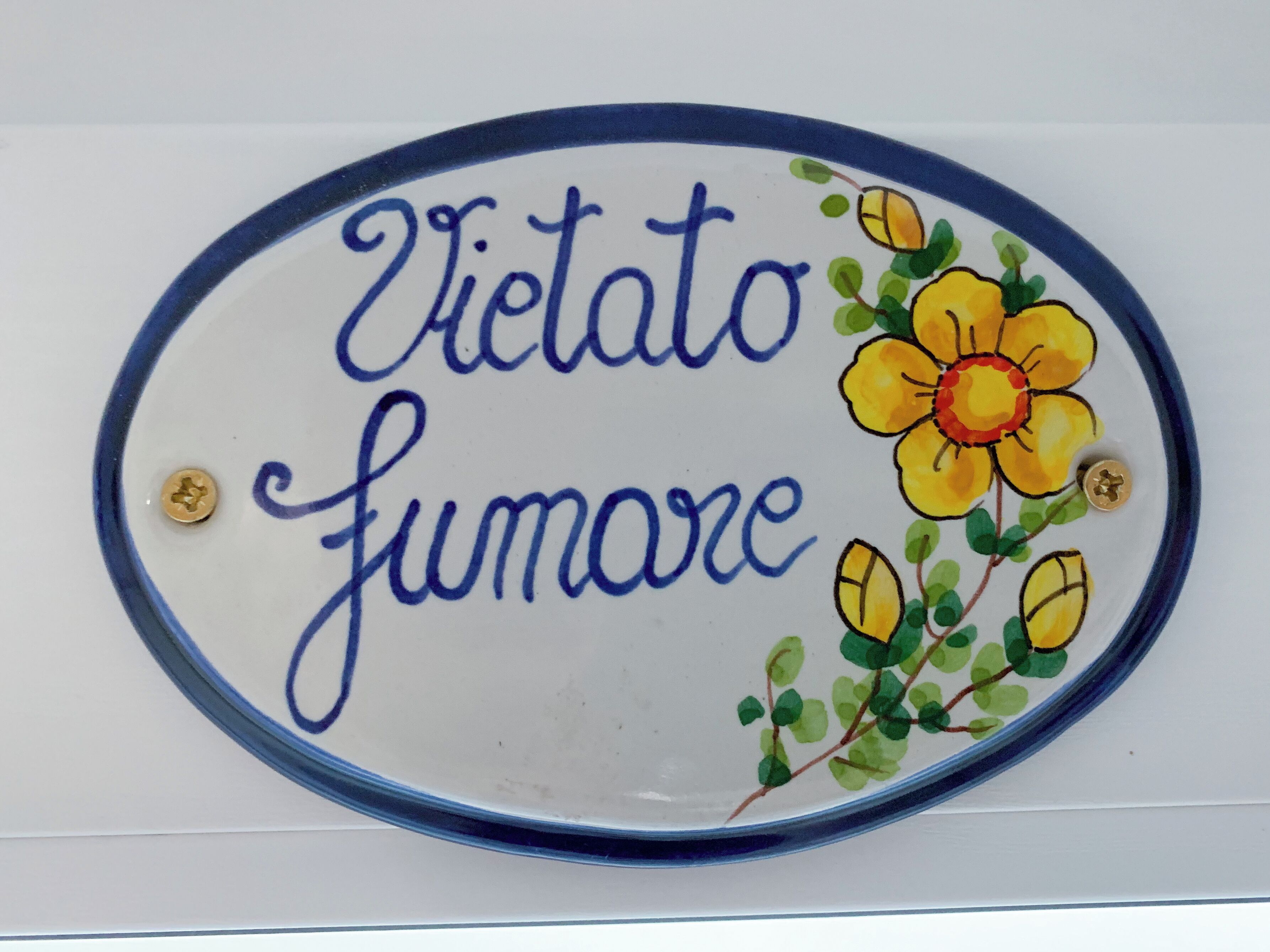 Um letreiro decorativo com o texto 'Vietato fumare' e uma ilustração de uma flor amarela.