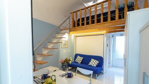 A modern living room with a blue sofa, a dining table with a vase of flowers, and a staircase leading to a loft area. - Freeholiday House in Amalfi Coast (Minori)