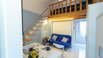 Una sala de estar moderna con un sofá azul, una mesa de comedor con un jarrón de flores y una escalera que lleva a un espacio tipo loft.