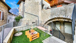 Un patio con un banco de madera, una mesa con una botella y un plato de uvas, y un muro de piedra.