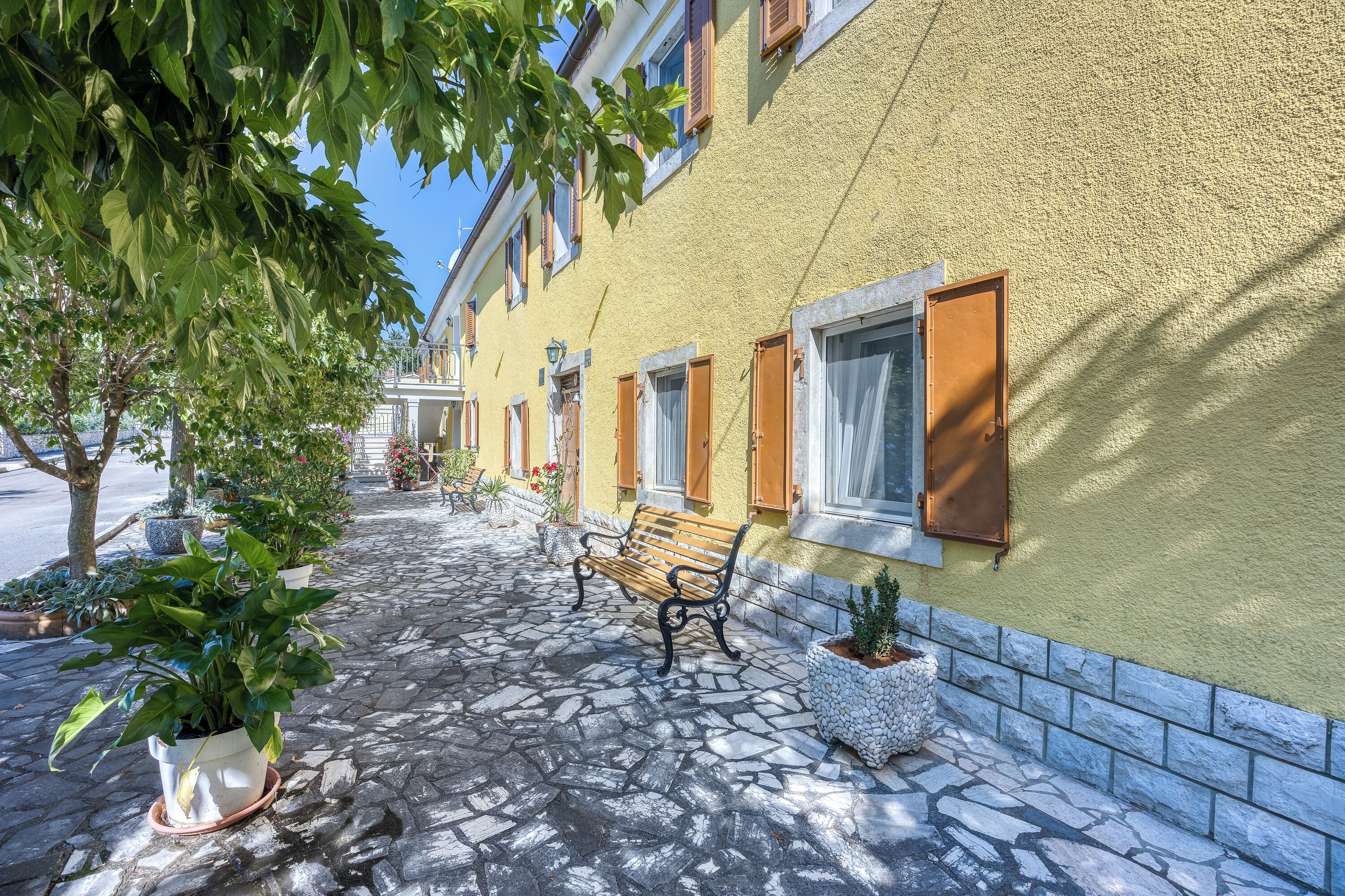 A cobblestone pathway with a bench, potted plants, and a building with a yellow facade and wooden shutters.