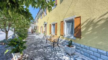 A cobblestone pathway with a bench, potted plants, and a building with a yellow facade and wooden shutters.