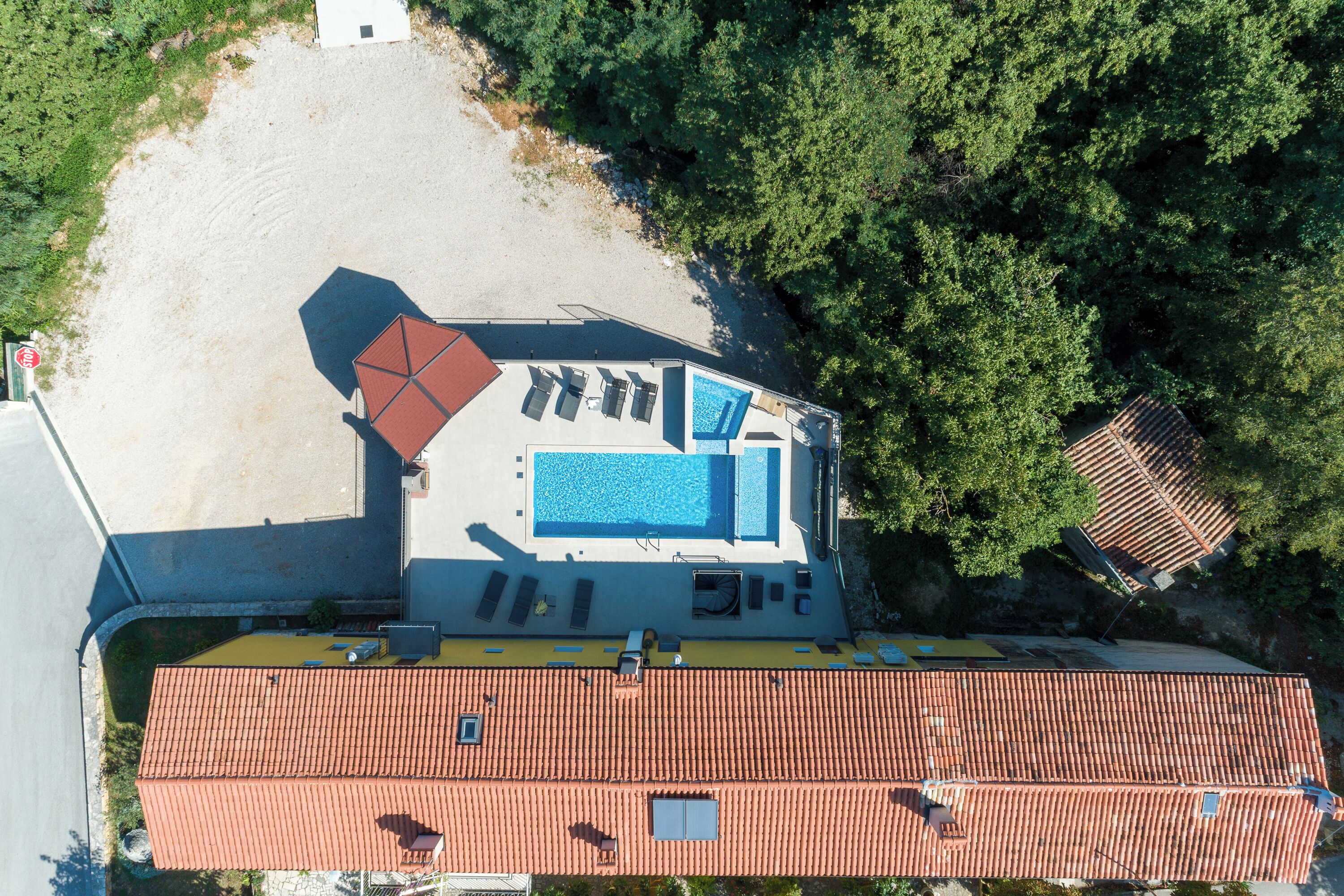 An aerial view of a property with a swimming pool, a red-roofed building, and a surrounding area with trees.