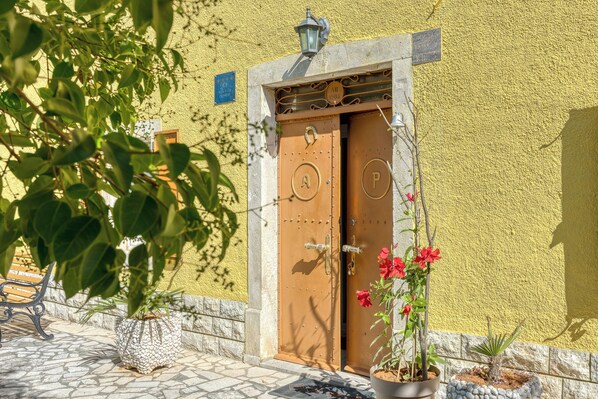 A building entrance with a yellow wall, a wooden door with metal handles, and a potted plant with red flowers.