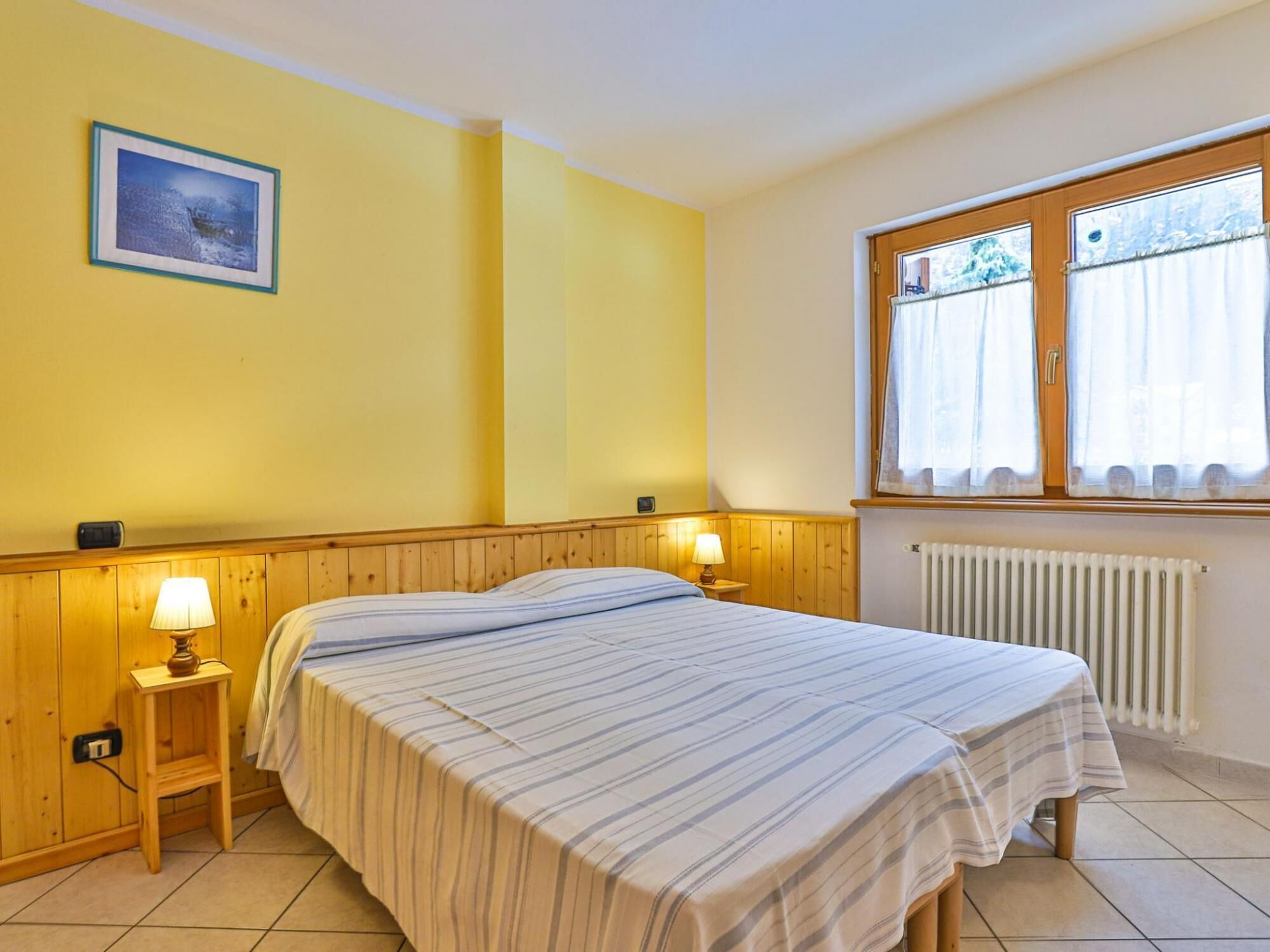 Une chambre d’hôtel avec un lit, des murs recouverts de lambris en bois, une fenêtre avec des rideaux, un radiateur et une petite table avec une lampe.