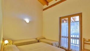 A room with two beds, a wooden bench, a table, and a sliding glass door leading to an outdoor area. - Apartment La Gran Becca - ID 3949 (Antey-Saint-Andre)