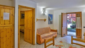 Una habitación con un armario de madera, un sofá, una cama con colcha estampada, una lámpara fijada a la pared y una puerta que da al exterior.