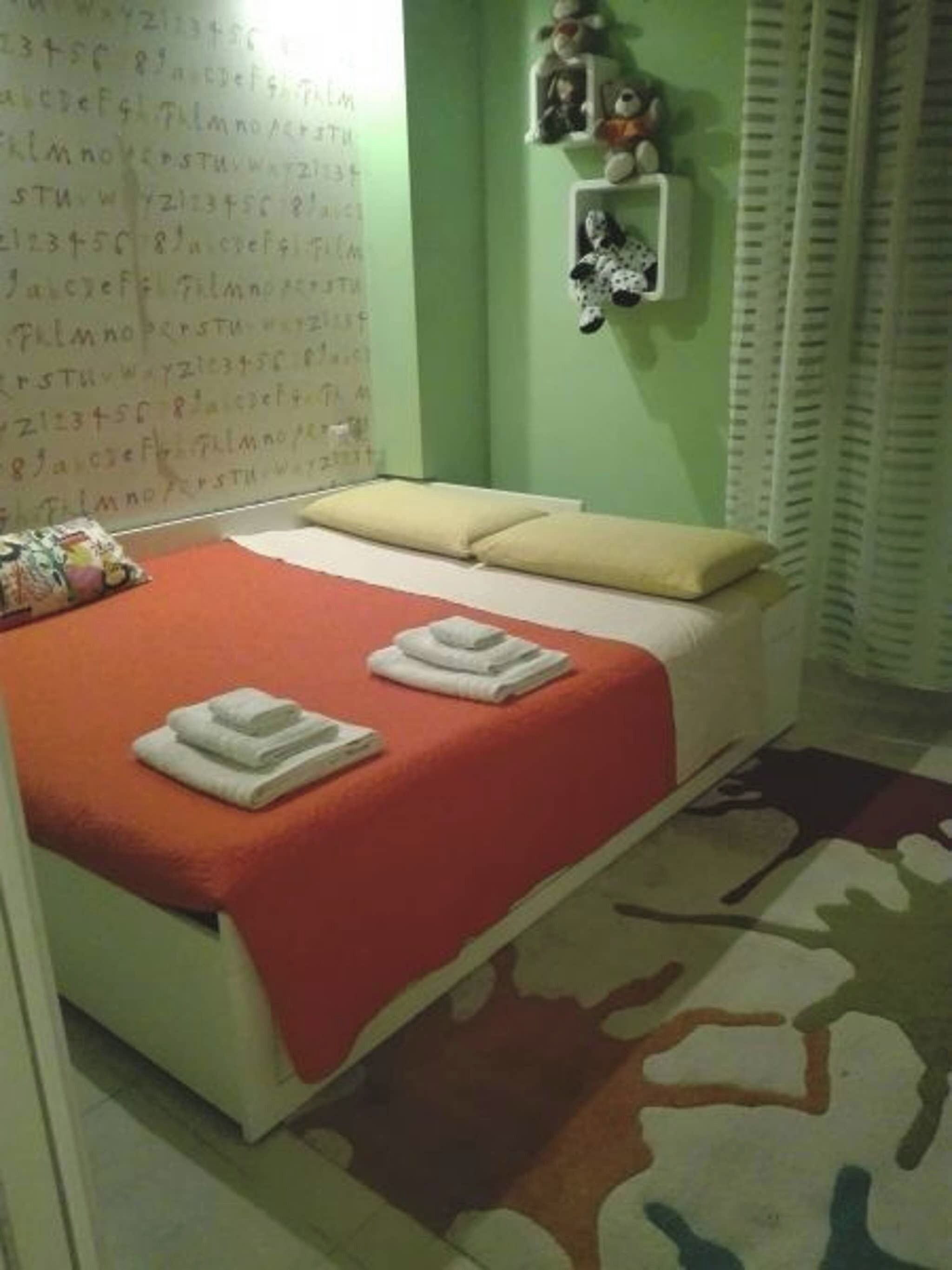 A bedroom with a bed, pillows, a wall with a letter chart, and a small shelf with decorative items.