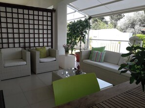 Uma área de estar moderna ao ar livre, com móveis de vime, uma mesa de vidro e plantas em vasos.