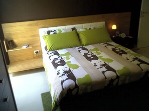Quarto com cabeceira de madeira, cama com colcha estampada, almofadas verdes, mesa de cabeceira com abajur e tomada.