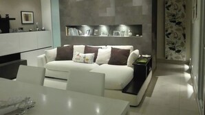 Una sala de estar moderna con un sofá blanco, una mesa de comedor y una zona de cocina con estantería empotrada.
