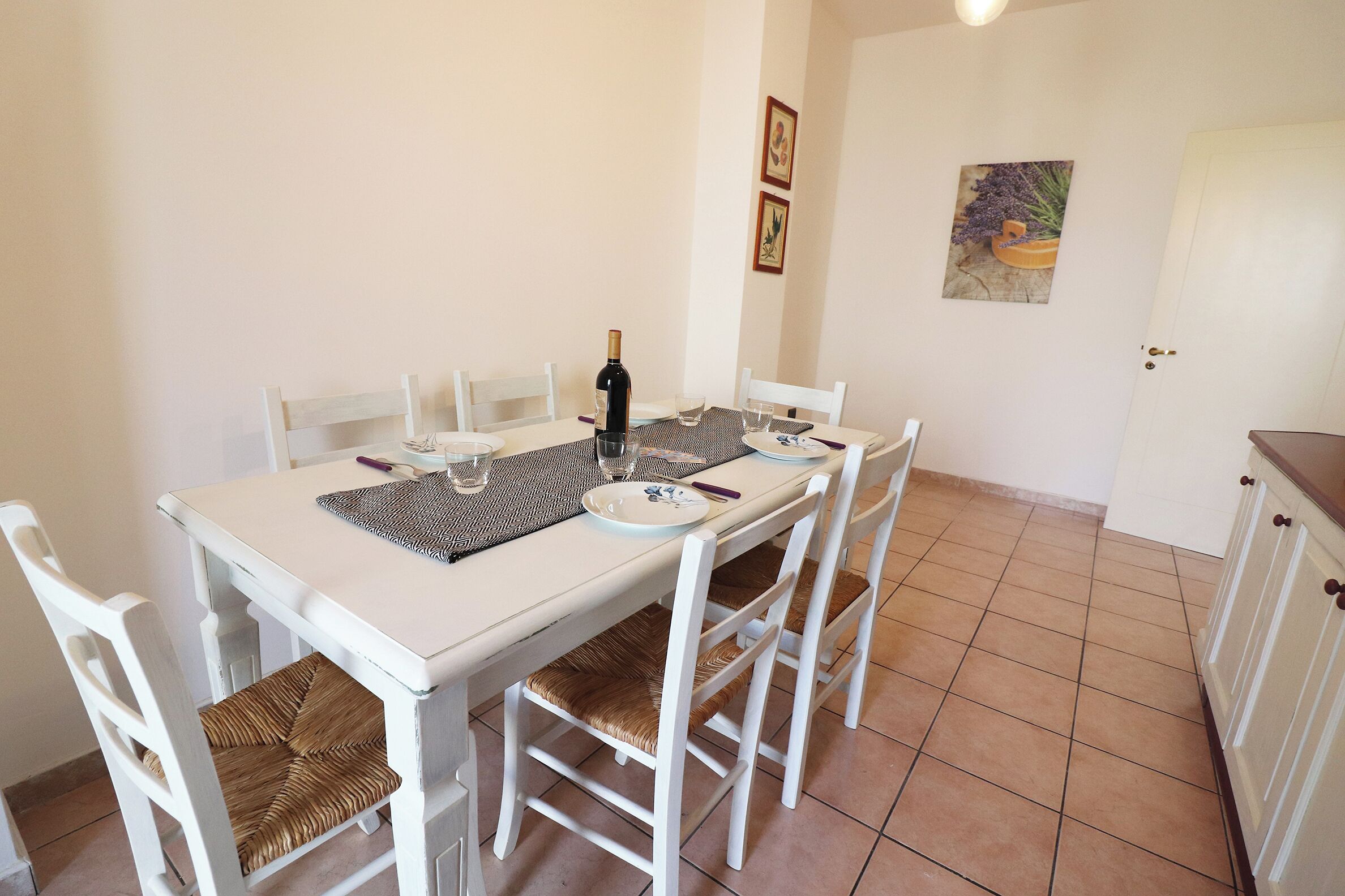 A dining area with a white table, four white chairs, and a bottle of wine.