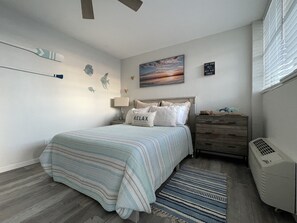 Ein Schlafzimmer mit Bett, Nachttisch, Kommode, Wandregal, Fenster mit Jalousien und Deckenventilator.