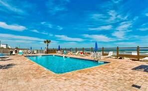 A rectangular swimming pool with a tiled deck, surrounded by lounge chairs and umbrellas, with a view of the ocean and a clear sky. - Steps to the Beach ! (Daytona Beach)