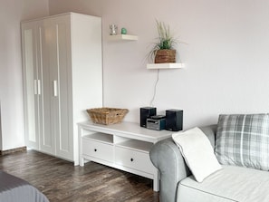 Un salon moderne avec une armoire blanche, une console blanche agrémentée d’un panier et d’enceintes, un canapé gris avec un coussin à carreaux et une plante posée sur une étagère murale.