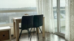 A dining area with a table and two chairs, a small table with a jar, and a view of a parking lot through a large window with sheer curtains. - FB06.05 - Modern studio apartment with a view from the 6th floor - Haus Frische (Cuxhaven)
