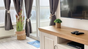 A modern room with a large TV, a wooden cabinet, a small plant, and a potted plant by the window. - FB06.05 - Modern studio apartment with a view from the 6th floor - Haus Frische (Cuxhaven)