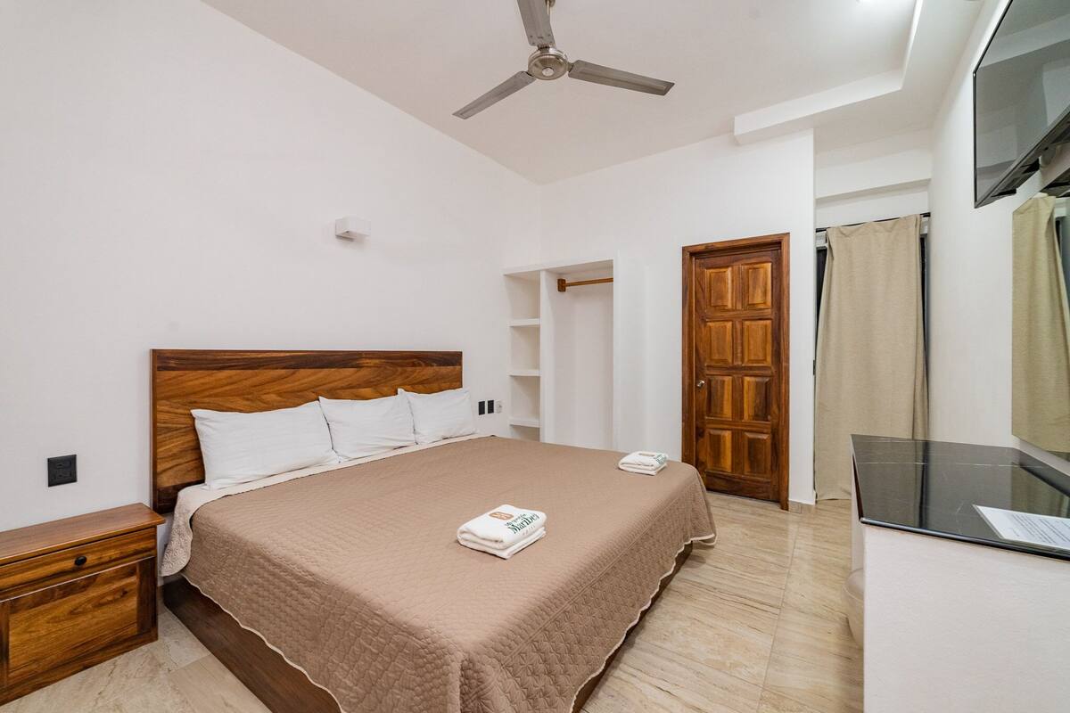 Un dormitorio con cabecera de madera, cama con colcha color beige, mesita de noche, ventilador de techo y televisor sobre un mueble bajo.
