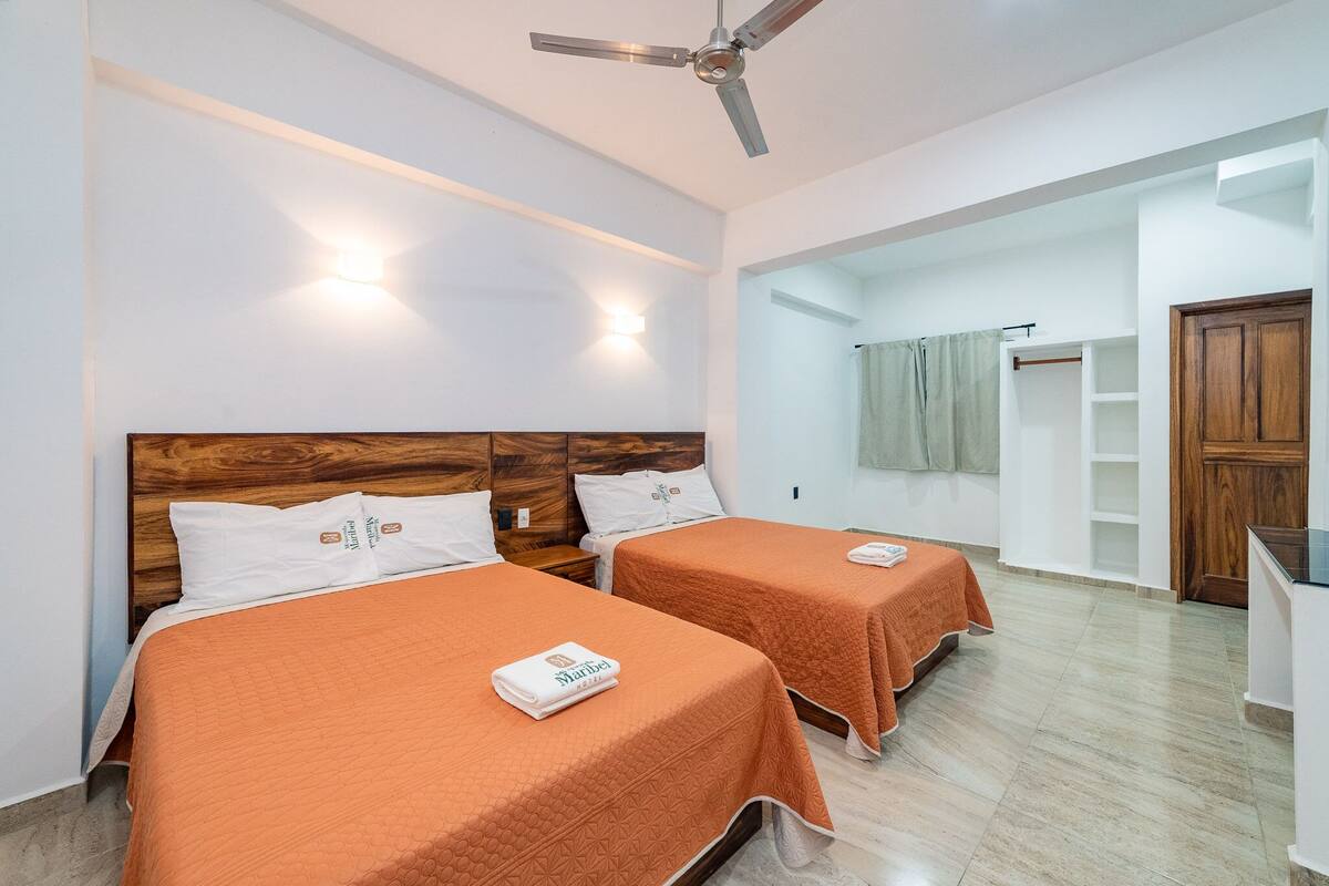 Habitación de hotel con dos camas, ventilador de techo, armario y baño visible a través de una puerta abierta.
