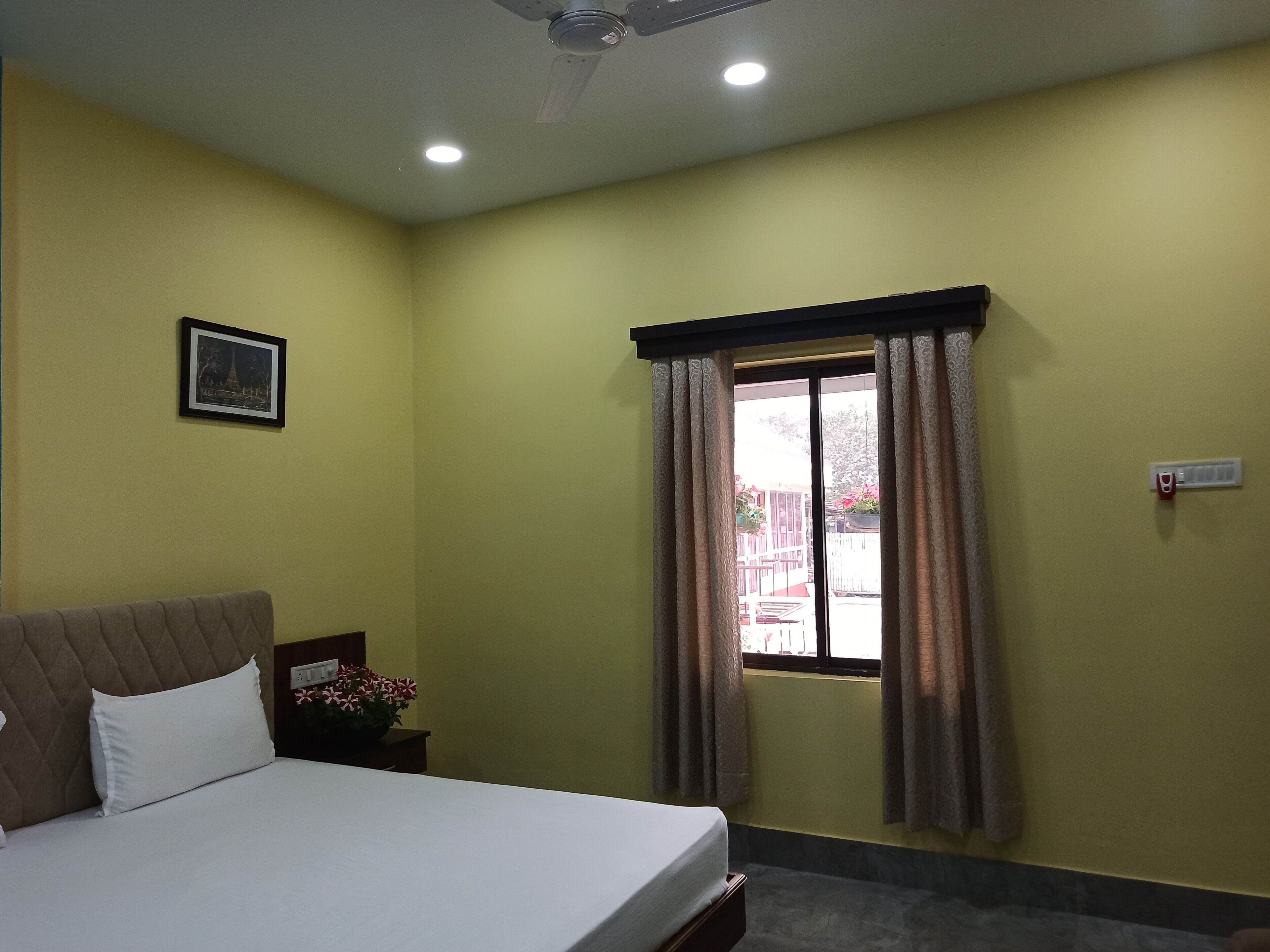 Una habitación de hotel con cama, mesita de noche, ventana con cortinas, ventilador de techo y una lámpara de pared.