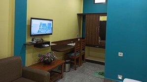 Una habitación pequeña con escritorio, televisor, silla, sofá, cesto de basura y ventana.