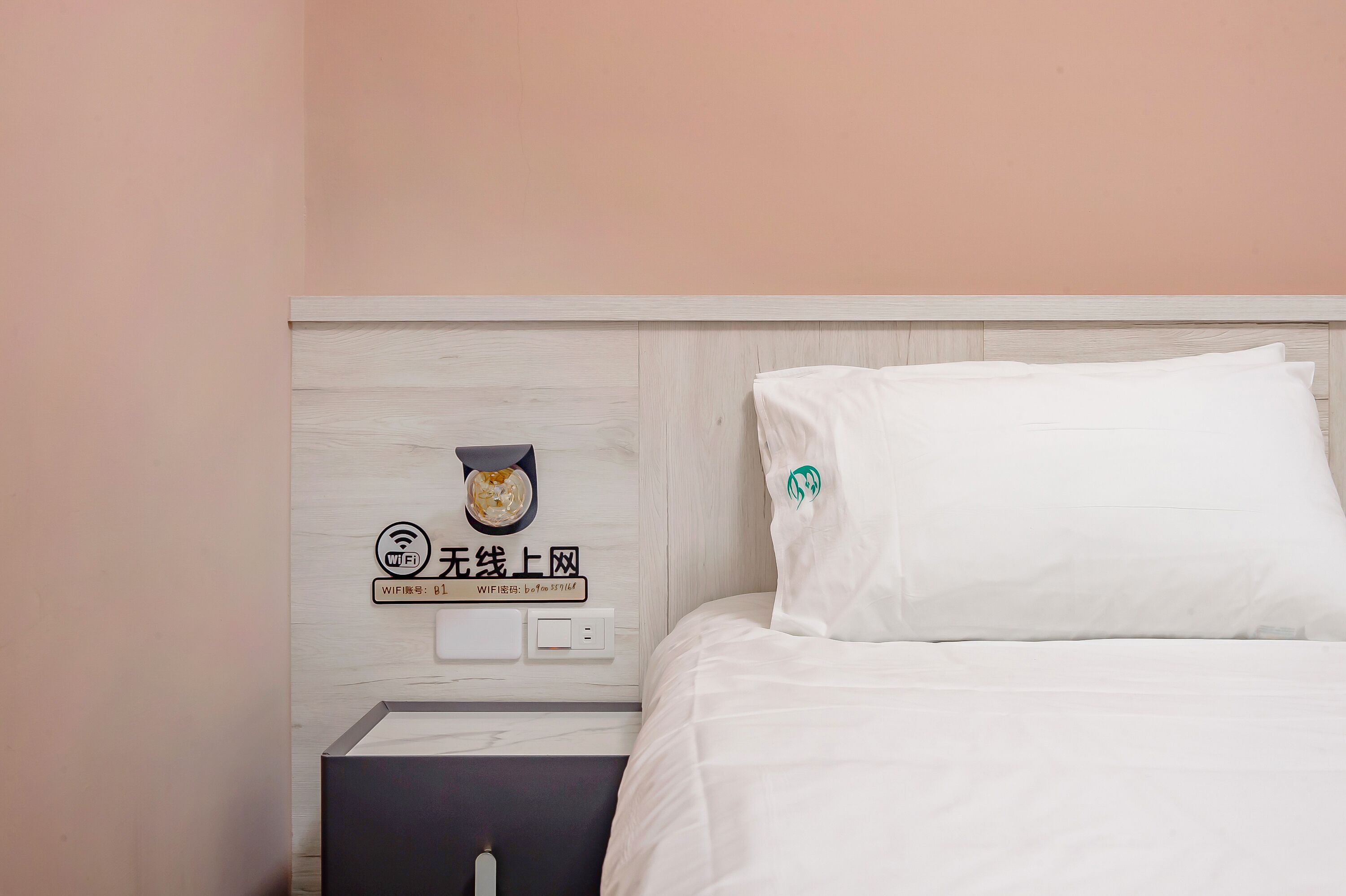 Un lit bien fait, avec une literie blanche et un oreiller orné d’un logo vert. Une petite table à la surface noire est visible à côté du lit.