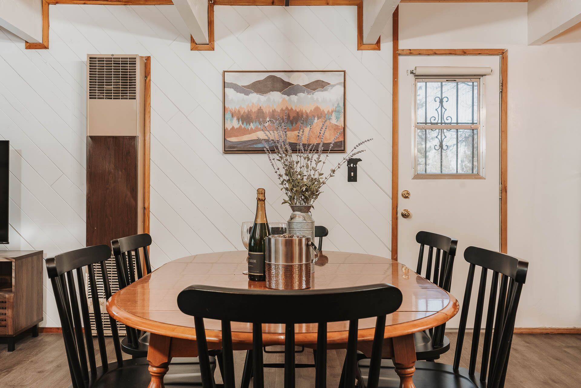 A dining area with a round wooden table, six black chairs, a painting of a mountain landscape, a door with a window, and a wall-mounted air vent.
