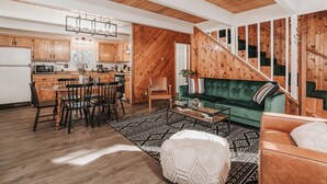 A spacious living area with a green sofa, wooden dining table, and chairs. A staircase with wooden railings leads to another level.