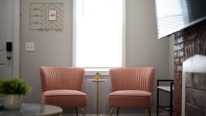 Ett rum med två rosa fåtöljer, ett litet bord, ett fönster med persienner, en dekorativ väggdekoration och en tegelvägg.