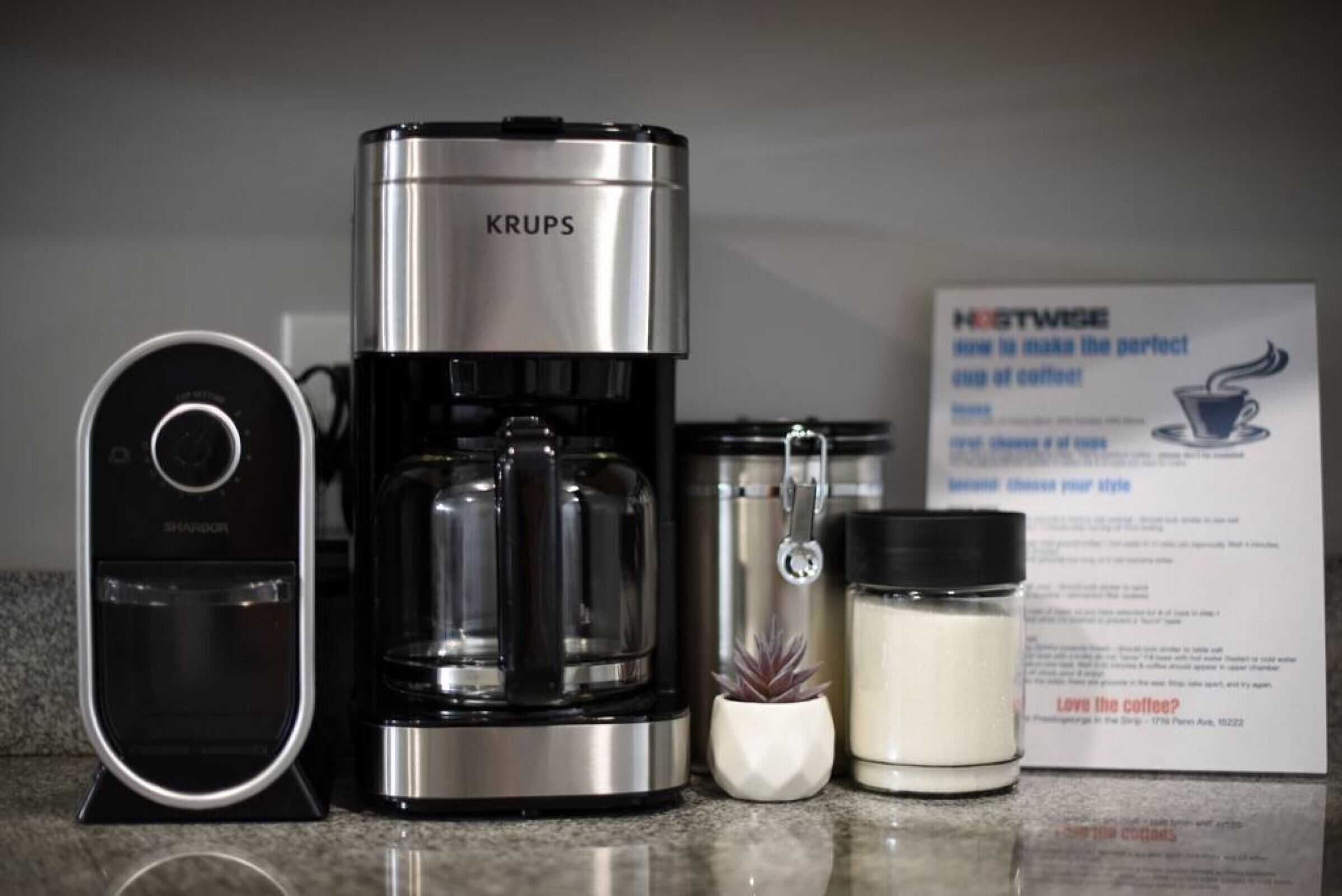 A Krups coffee maker, a ShaverPro device, a sign with coffee-related text, and a small potted plant.