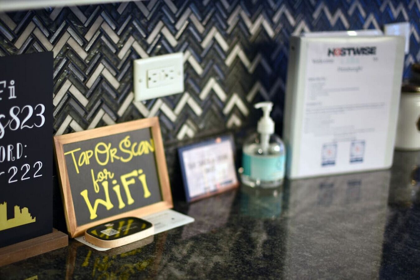 A hotel reception area with a chalkboard sign advertising a top scan for WiFi, a digital screen, a Nestweave sign, and a hand sanitizer dispenser.