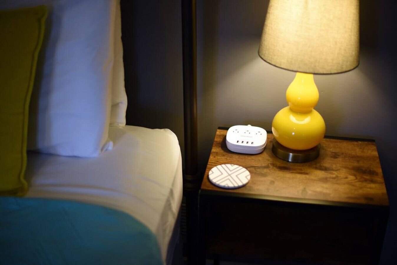 A neatly made bed with white linens and a yellow pillow, a bedside table with a yellow lamp, a power strip, and a decorative coaster.