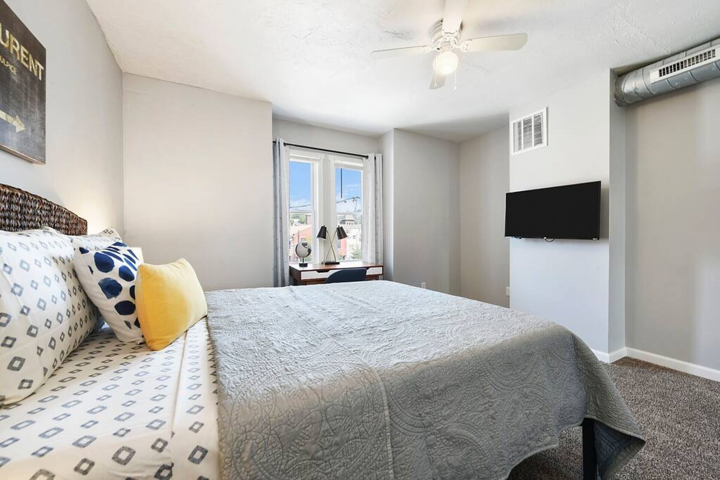 A bedroom with a bed, a desk, a chair, a ceiling fan, a TV, and a window.