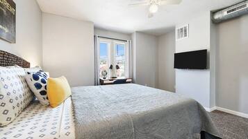 A bedroom with a bed, a desk, a chair, a ceiling fan, a TV, and a window.